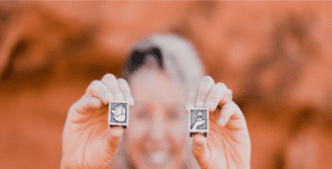 A person is holding two small, square tiles with illustrations—one showcasing a thumbs-up and the other a pointing dog—capturing the essence of love language. The blurred background bathes the scene in an earthy, red tone.