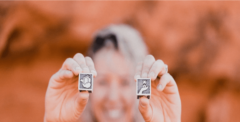 A person is holding two small, square tiles with illustrations—one showcasing a thumbs-up and the other a pointing dog—capturing the essence of love language. The blurred background bathes the scene in an earthy, red tone.