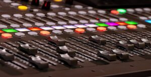 Close-up view of a professional audio mixing console with numerous sliders, buttons, and lights in various colors. The sliders and buttons are meticulously arranged in rows, illustrating the science of sound as they adjust levels and effects for precise sound mixing.