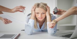 A frustrated woman sits at a desk, resting her head in her hands. Surrounding her, four hands hold a phone, a tablet, a watch, and papers—an overwhelming scene needing stress management. A laptop and a notebook are visible on the desk.