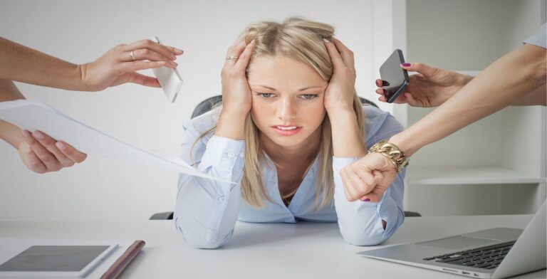 A frustrated woman sits at a desk, resting her head in her hands. Surrounding her, four hands hold a phone, a tablet, a watch, and papers—an overwhelming scene needing stress management. A laptop and a notebook are visible on the desk.
