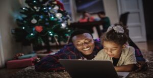 A father and daughter lie on the floor, smiling while watching a Christmas stream on their laptop. A decorated tree with lights and presents is in the background, along with two people sitting at a table. The room is warmly lit, creating a cozy atmosphere.