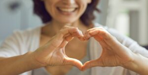 A person smiles while forming a heart shape with their hands in front of them, embodying the spirit of holiday appreciation. The background is softly blurred, and the lighting is warm and gentle.
