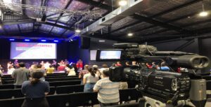A camera streams a church service where the audience stands, with a stage in the background. The screen displays "Tithes & Offerings" in red text, ensuring visibility. The ceiling is industrial with visible beams.