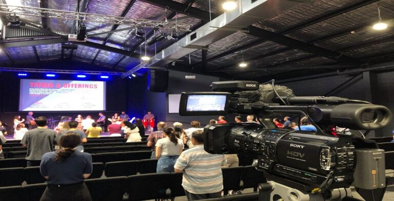 A camera streams a church service where the audience stands, with a stage in the background. The screen displays "Tithes & Offerings" in red text, ensuring visibility. The ceiling is industrial with visible beams.