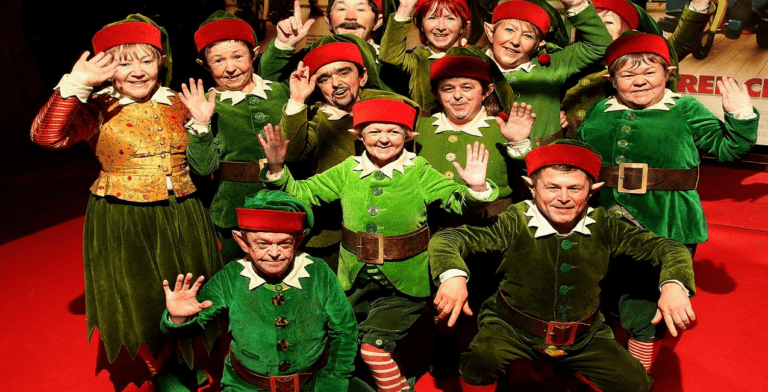 A group of people dressed as elves pose and wave enthusiastically, their festive green costumes with red hats and belts adding to the Christmas spirit. The background features holiday-themed decorations, reminiscent of a holiday event at a thriving church tech gathering. They stand proudly on a red carpet.