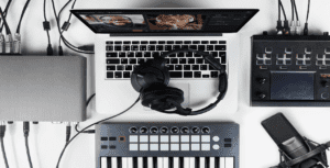 A music production setup featuring a laptop displaying a video call, streaming equipment like headphones on the keyboard, audio control devices, and a microphone. Various cables are connected, ensuring clarity in this professional studio environment.