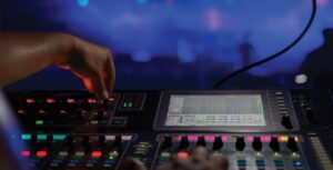 A person expertly adjusts settings on an audio mixing console, the brightly lit buttons and sliders gleaming under the stage lights. The background, a blur of blue and purple hues, hints at a live streaming event where top audio mixing tips are showcased in real-time.
