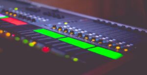 Close-up of an audio mixing console with various buttons and sliders, inviting tech volunteers to engage in this tech-tonic shift. Some buttons are illuminated in red and green, indicating active channels. The dim lighting highlights the equipment’s sleek design and colorful interface.