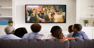 A diverse group of five engaged viewers sits on a couch in a bright living room, streaming a concert on the wall-mounted television. They appear relaxed amid the room's plants and decorative items on shelves.