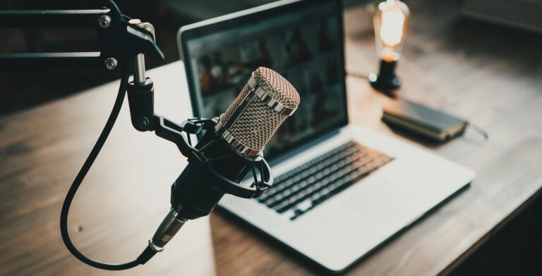 A close-up of a microphone on a stand hints at an upcoming podcast, set in front of an open laptop on a wooden desk. The screen displays a blurred image, while a small notebook beside it awaits simple steps to be jotted down under the warm glow of an Edison bulb lamp.