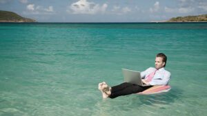 A man in business attire relaxes on a float in clear blue water, live streaming from his laptop. The background shows a sunny beach and clear summer skies.