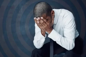 A man in a white dress shirt and black pants sits with his head in his hands, appearing distressed, as if pondering past mistakes. The background features circular patterns reminiscent of stained glass windows in churches.