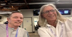Two people are smiling for a selfie indoors, both donning lanyards; one reads "MENTOR." The setting resembles an office or conference space with exposed ceiling elements and wall screens. Their laughter fills the room, creating a legacy of joy captured forever in this snapshot.