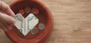 A hand holds a fifty-dollar bill folded into a heart shape above a wooden bowl of assorted coins, illustrating Church stewardship on a light brown surface.