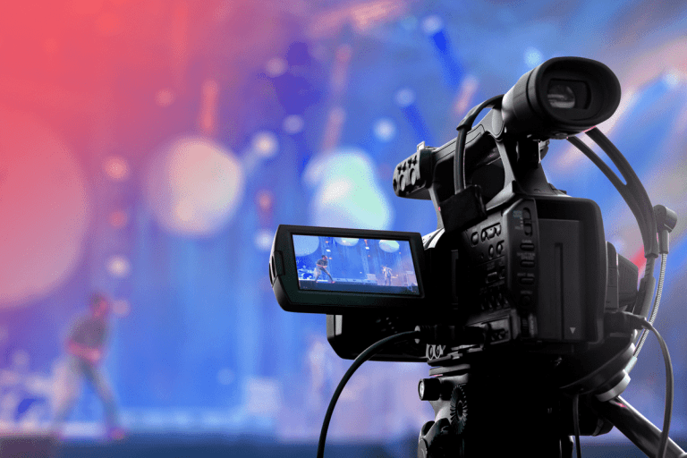 A close-up of a video camera recording a live event on stage with colorful, blurred lights and a person performing in the background—perfect for streaming video for churches.