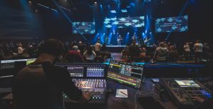A sound engineer operates a control board at the back of a dimly lit venue while a band performs on stage under blue lights, with an audience watching—mirroring the setup often used in church live streaming for enhanced worship experiences.