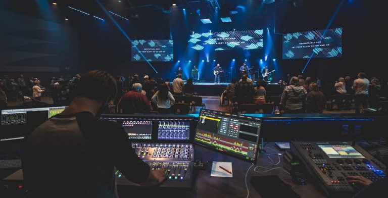 A sound engineer operates a control board at the back of a dimly lit venue while a band performs on stage under blue lights, with an audience watching—mirroring the setup often used in church live streaming for enhanced worship experiences.